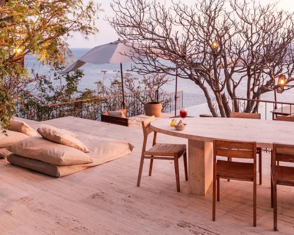 Terrasse en bois avec table à manger et coussins de sol face à la mer.
