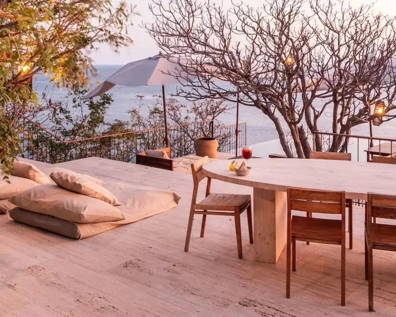 Terrasse en bois avec table à manger et coussins de sol face à la mer.