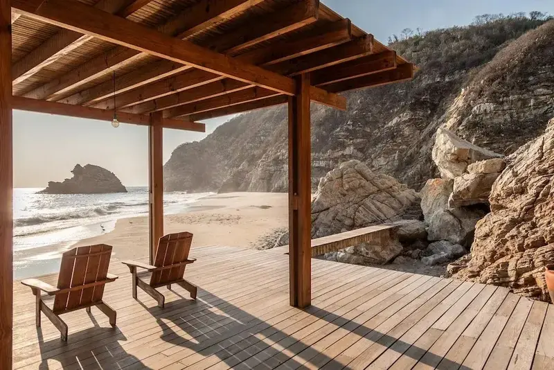 Deux fauteuils en bois sont placés sur une terrasse en bois donnant sur une plage de sable et la mer.