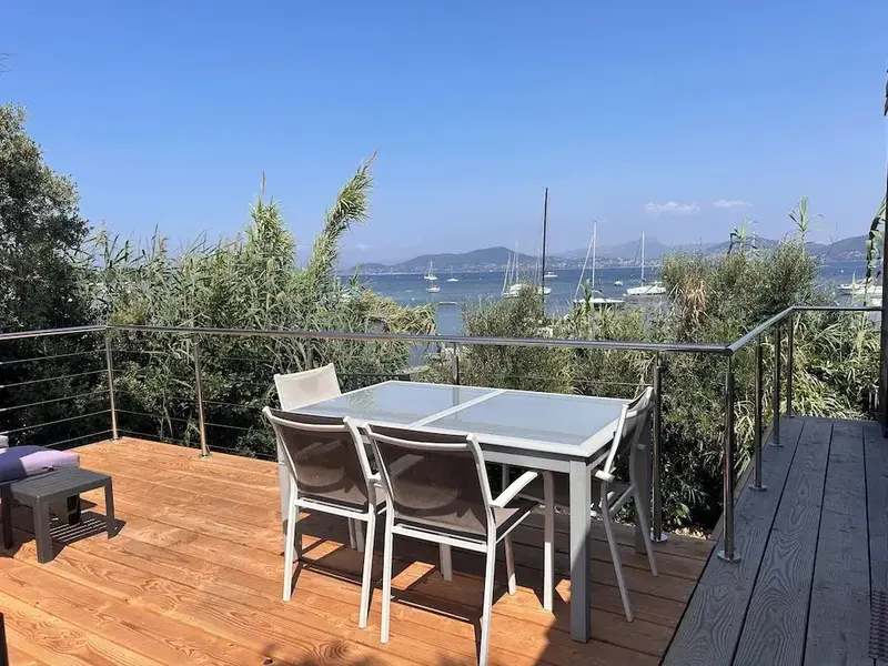 Une table et des chaises en métal sur une terrasse en bois avec vue sur la mer et des bateaux.