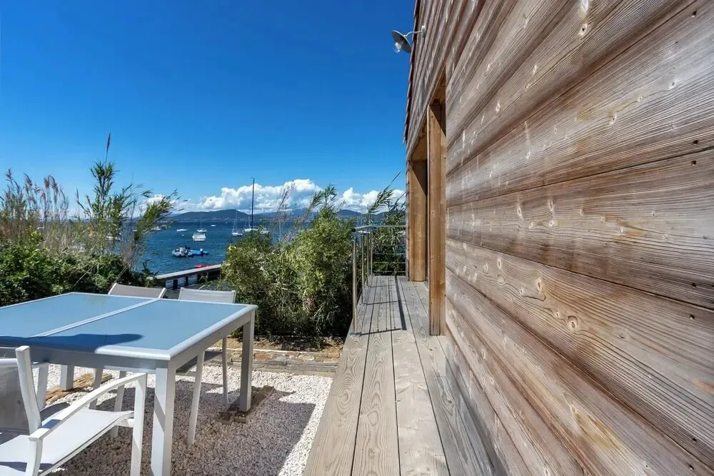 Une table et des chaises en métal se trouvent sur une terrasse en bois avec vue sur la mer et les bateaux.