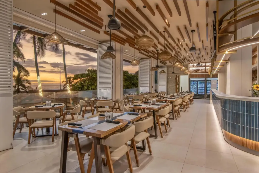 Une salle de restaurant lumineuse avec des tables dressées face à un coucher de soleil sur la mer.