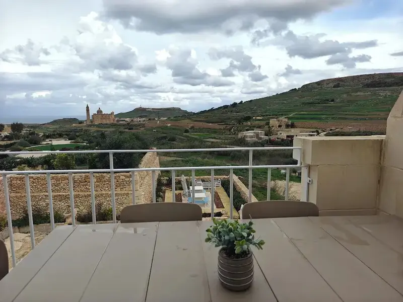 Terrasse avec table en bois donnant sur un paysage vallonné et une église au loin.