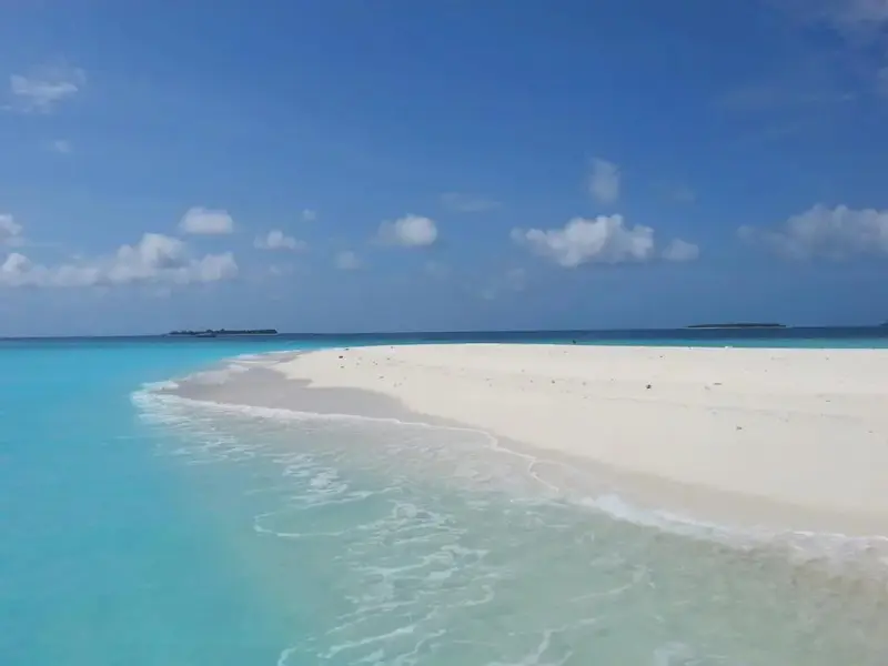 Plage de sable blanc bordée d'eau turquoise sous un ciel bleu nuageux.