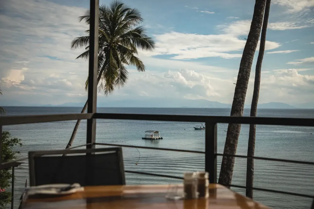 Une table en bois est mise en scène devant une vue sur la mer et des palmiers.