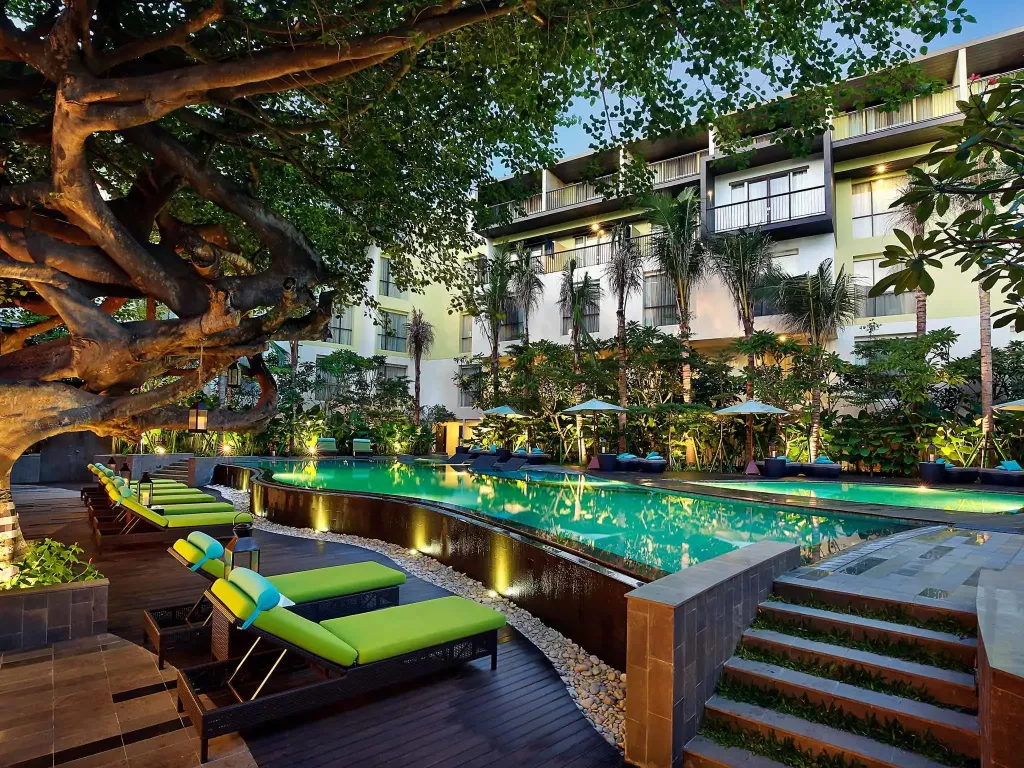 Transats verts alignés au bord d'une piscine illuminée, avec un bâtiment moderne en arrière-plan.