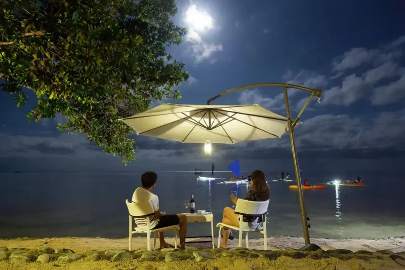 Une table est dressée sur le sable sous un parasol, face à la mer illuminée par la lune.
