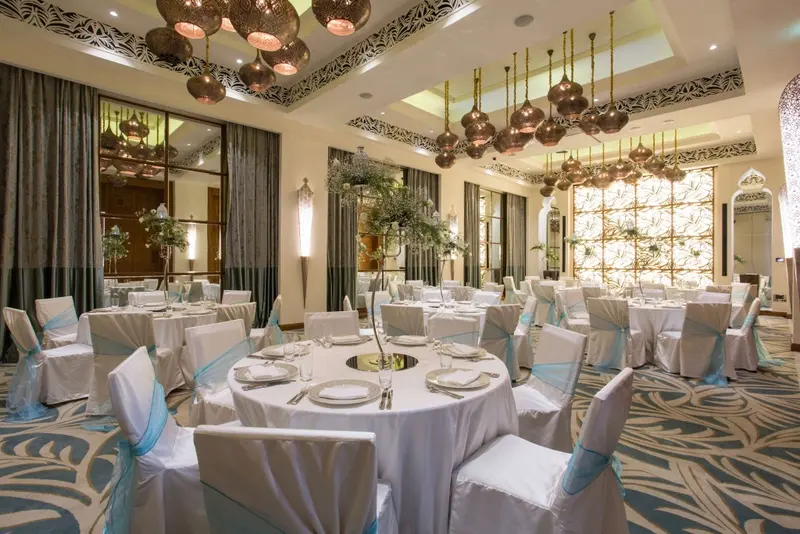Une salle lumineuse est aménagée avec des tables rondes nappées et des chaises habillées.