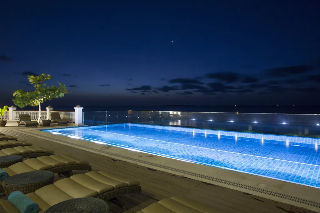 Piscine illuminée avec transats et vue sur la mer nocturne.