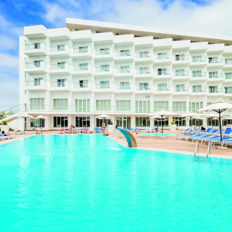 Une piscine turquoise bordée de transats blancs devant un bâtiment moderne.