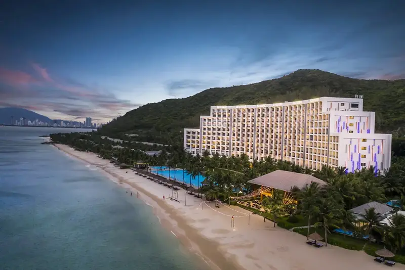 Vue aérienne d'un grand bâtiment blanc bordant une plage de sable fin et une mer turquoise.