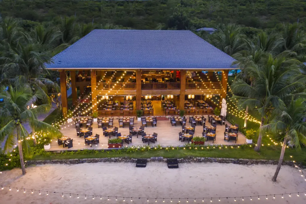 Vue aérienne d'un bâtiment éclairé avec une terrasse et des tables de repas en bord de mer.