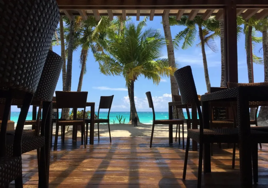 Terrasse ombragée avec tables et chaises donnant sur une plage de sable blanc et la mer turquoise.