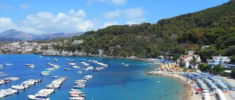 Une plage de sable bordée de transats bleus et de bateaux amarrés dans une baie turquoise.