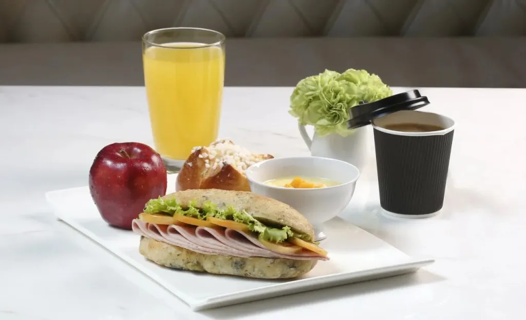 Un petit-déjeuner composé d'un sandwich, d'un jus d'orange, d'une viennoiserie, d'un fruit et d'une boisson chaude est présenté sur un plateau.