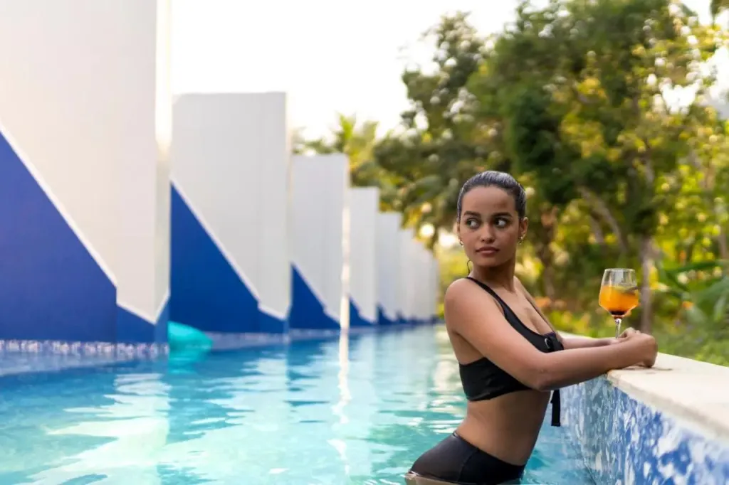 Une femme se tient au bord d'une piscine turquoise, un verre à la main.