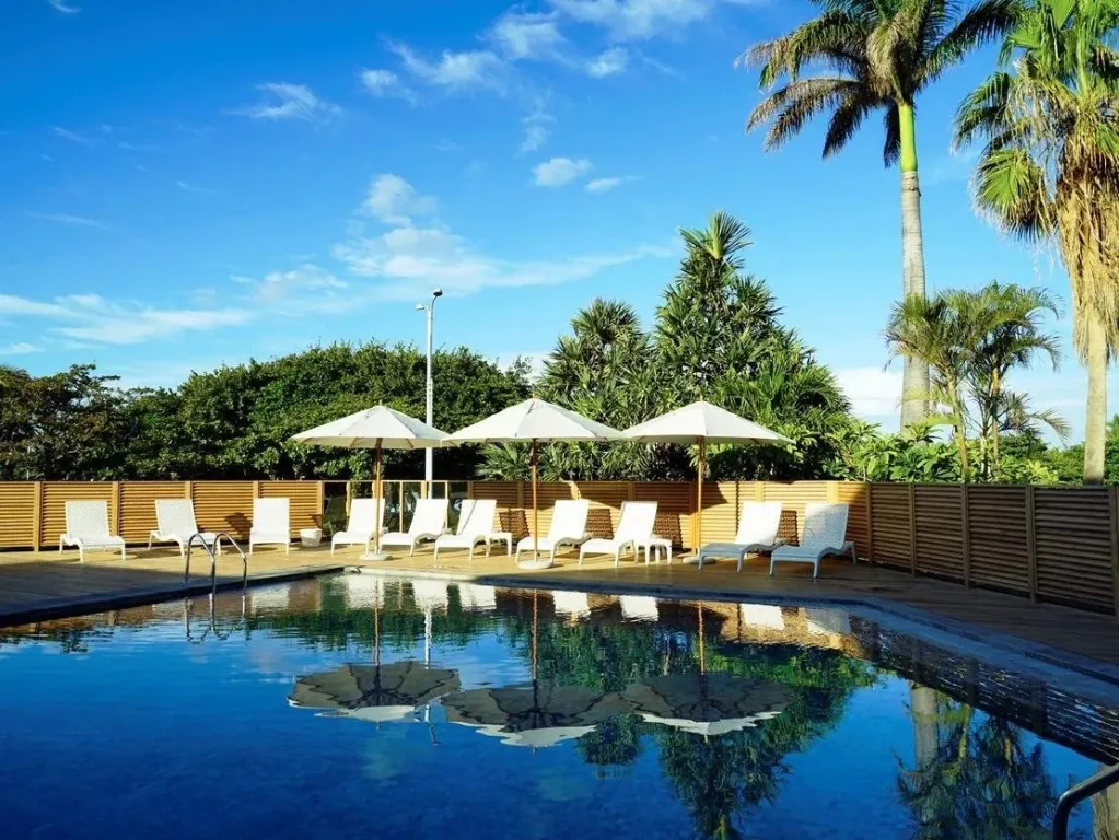 Transats blancs disposés au bord d'une piscine rectangulaire sous des parasols.