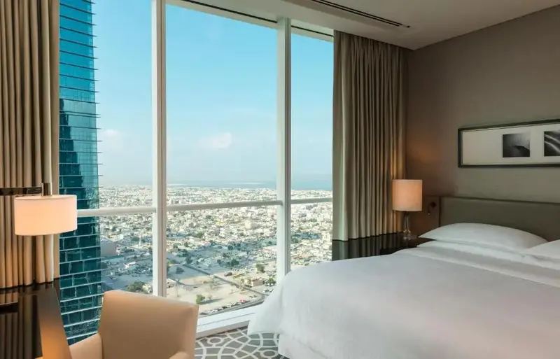 Une chambre lumineuse avec un grand lit face à une baie vitrée offrant une vue sur la ville et la mer.