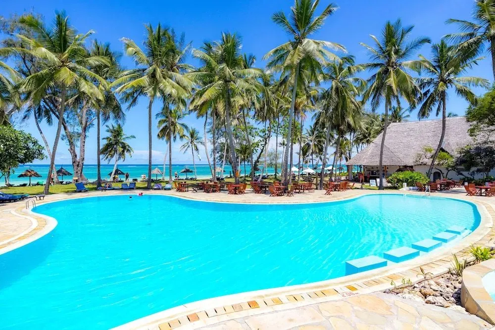 Une piscine turquoise bordée de palmiers et de transats, avec une plage en arrière-plan.