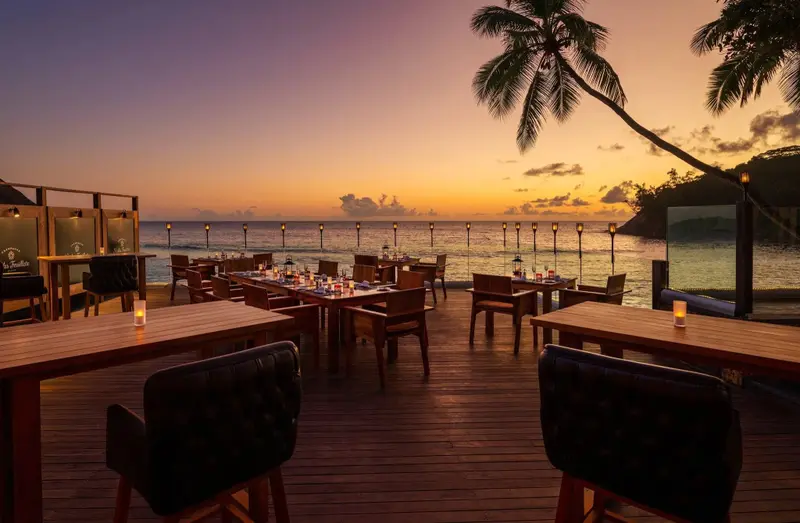 Tables de restaurant en bois disposées sur une terrasse face à la mer au coucher du soleil.