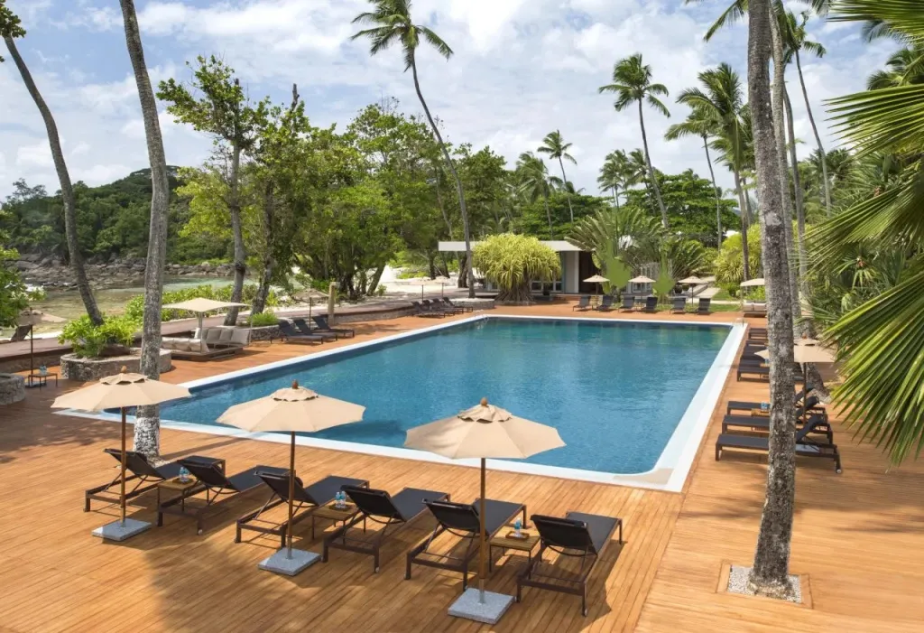 Une piscine rectangulaire bordée de transats sous des parasols, entourée de végétation tropicale.