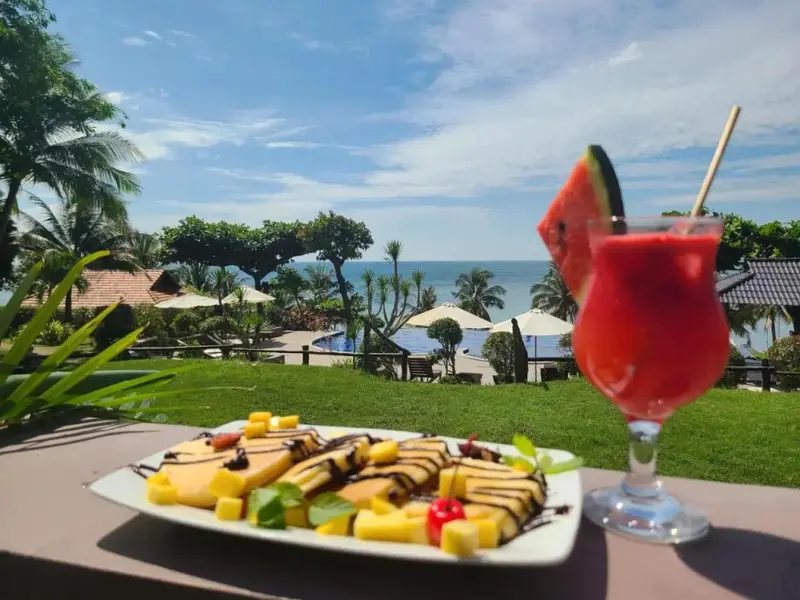 Un plat de crêpes aux fruits est présenté au premier plan, avec une vue sur la mer et la végétation luxuriante en arrière-plan.