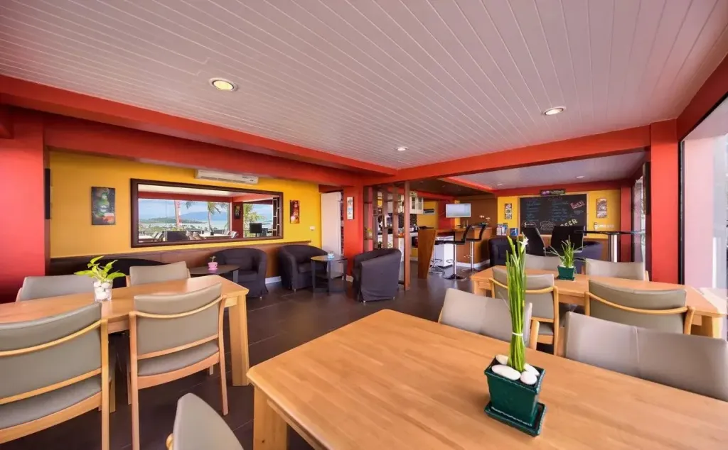 Une salle lumineuse avec des tables et des chaises, décorée de couleurs vives et donnant sur un paysage marin.