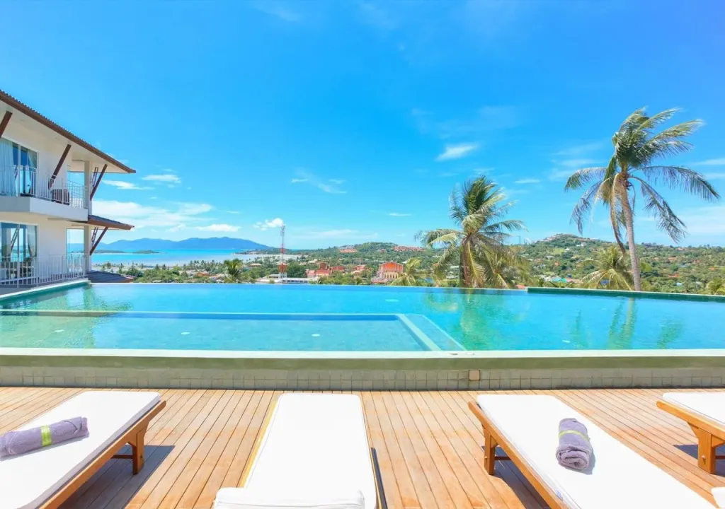 Transats blancs alignés au bord d'une piscine à débordement avec vue sur la mer et la végétation.