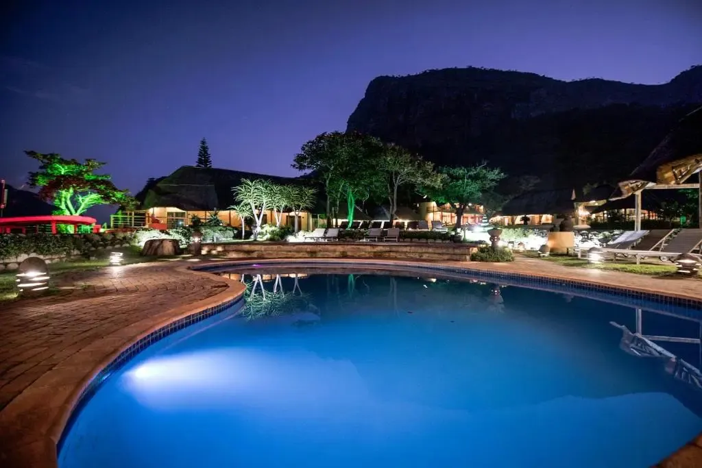 Piscine illuminée au premier plan avec un bâtiment éclairé en arrière-plan et une montagne sombre.