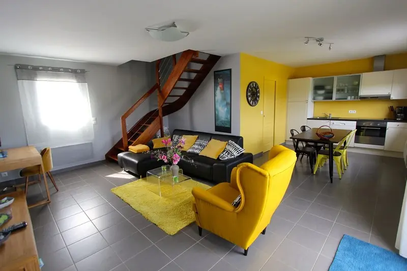 Un espace de vie lumineux avec un canapé noir, des fauteuils jaunes et une cuisine ouverte.