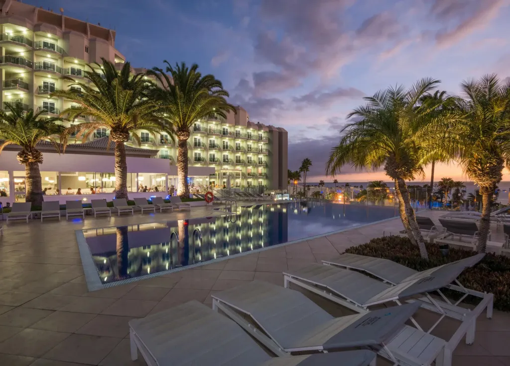 Une piscine bordée de transats devant un bâtiment illuminé au crépuscule.