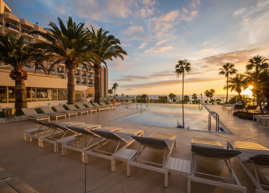 Transats blancs alignés au bord d'une piscine avec un bâtiment en arrière-plan et un coucher de soleil.