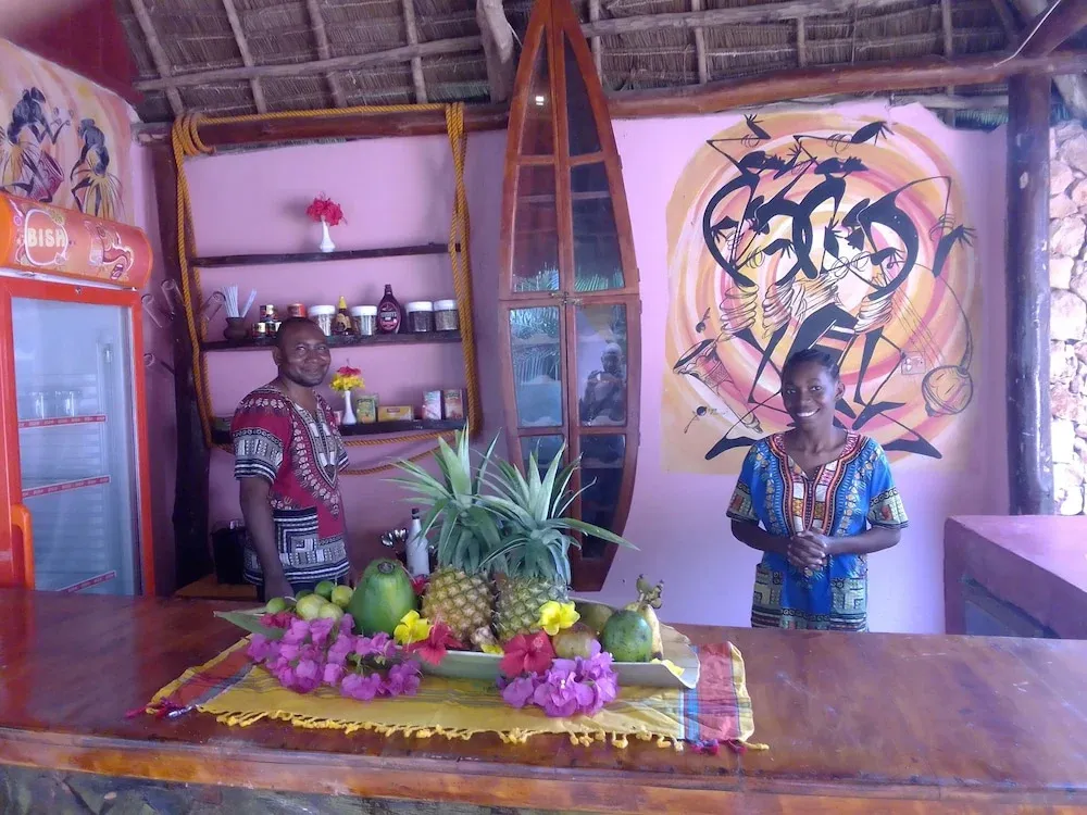 Un comptoir de bar en bois présente des fruits frais et des fleurs tropicales devant deux personnes souriantes.