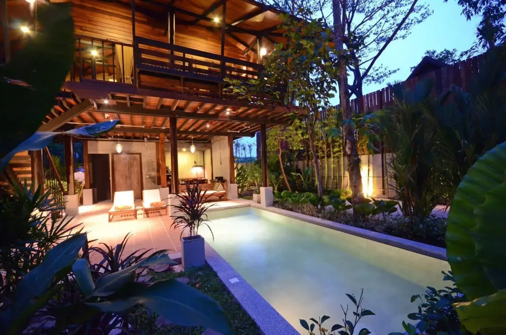 Une piscine rectangulaire bordée de végétation luxuriante, avec des transats et un espace couvert éclairé.