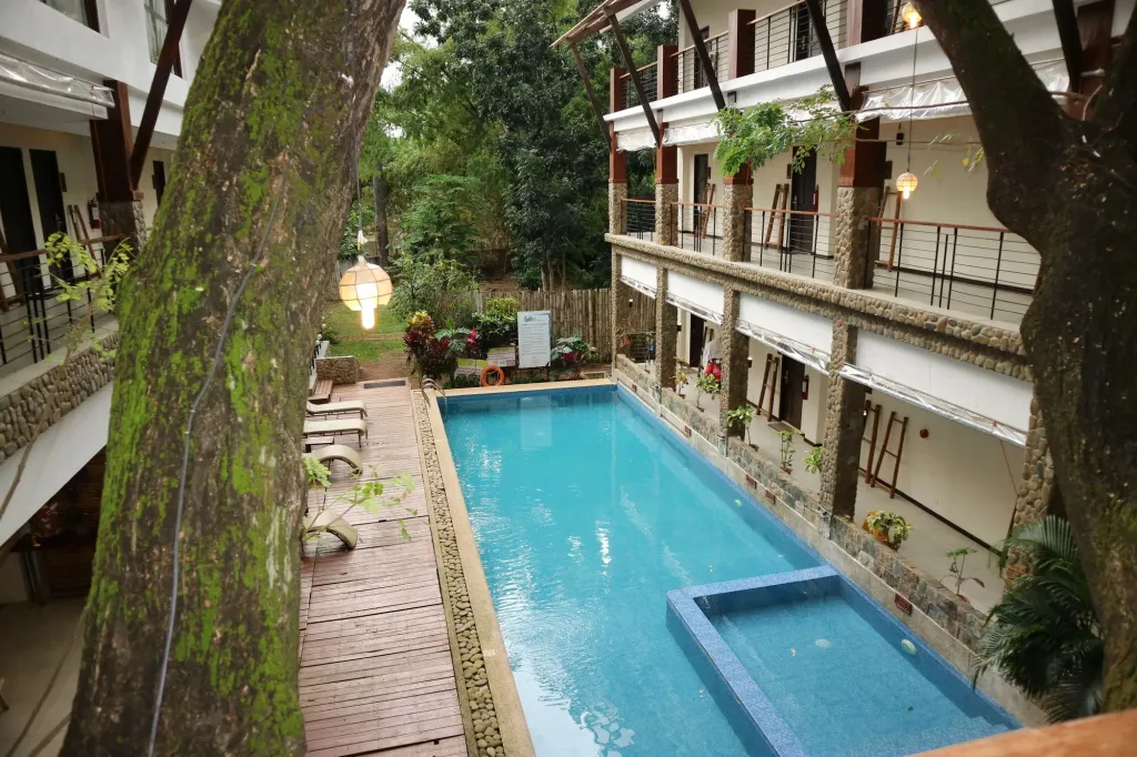 Une piscine rectangulaire est entourée d'une terrasse en bois et de bâtiments à balcons.