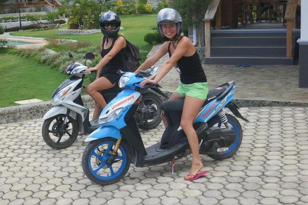 Deux femmes souriantes sont assises sur des scooters devant un bâtiment.