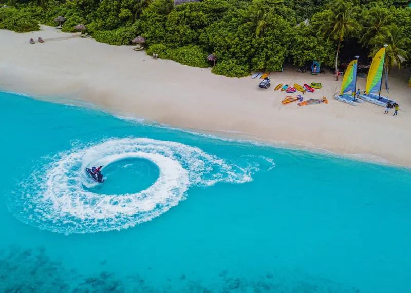 Une personne conduit un jet-ski sur une eau turquoise le long d'une plage de sable blanc.