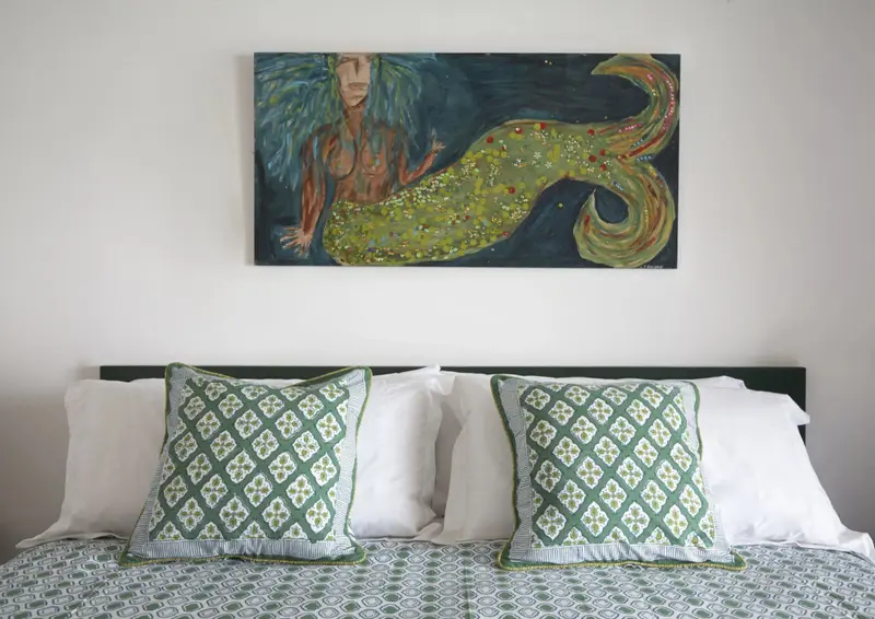 Un lit est fait avec des oreillers blancs et des coussins verts devant une peinture représentant une sirène.