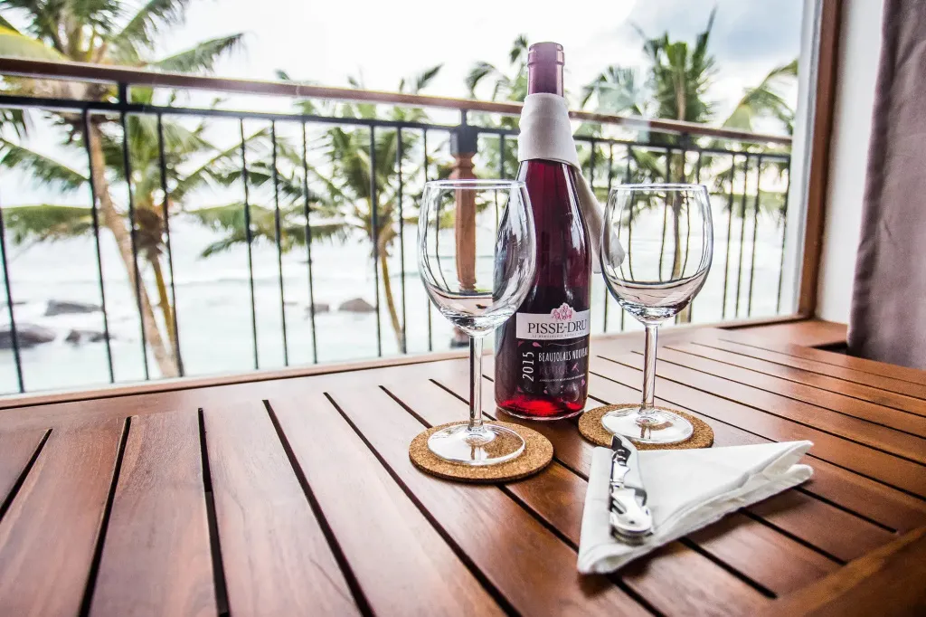 Une bouteille de vin et deux verres sur une terrasse en bois avec vue sur la mer et la végétation.