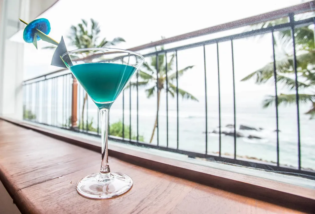 Un cocktail bleu est servi sur une rambarde en bois avec vue sur la mer et les palmiers.