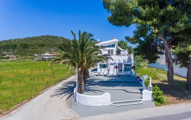 Bâtiment blanc à deux étages avec des palmiers devant et une colline verdoyante en arrière-plan.