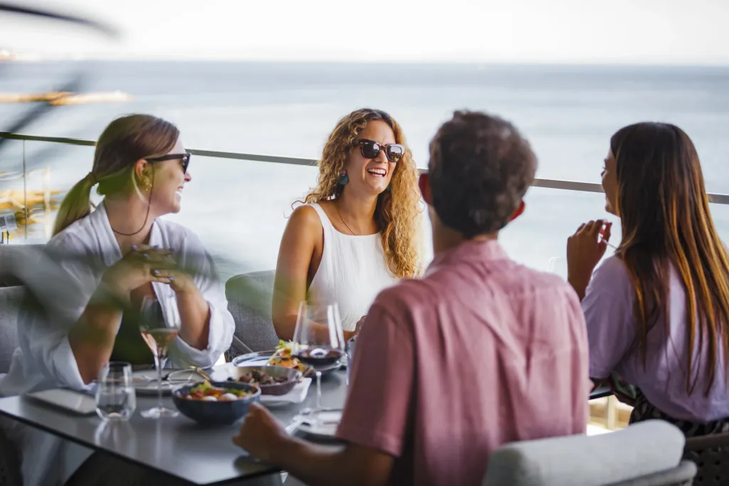 Des convives discutent autour d'une table en extérieur avec vue sur la mer.