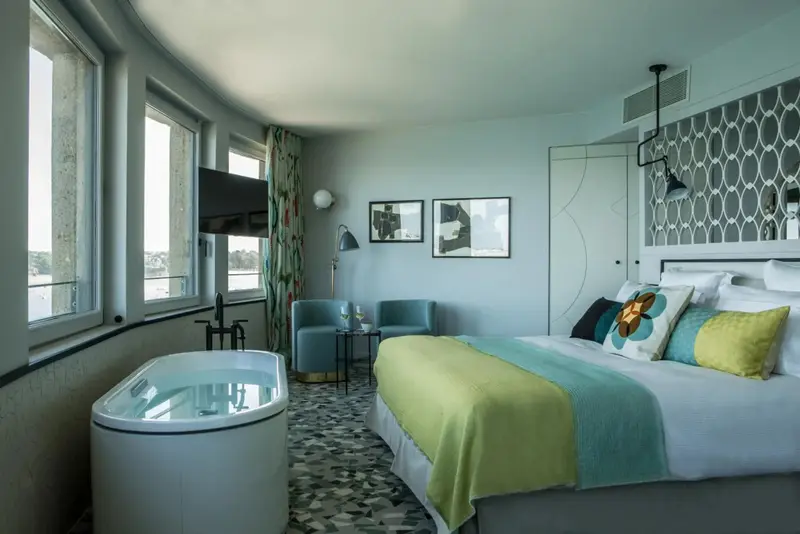 Une chambre lumineuse avec un lit recouvert d'une couverture verte et turquoise, et une baignoire blanche près des fenêtres.