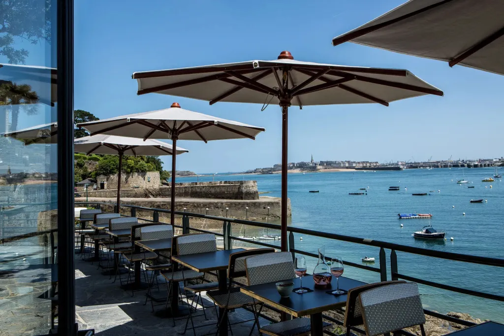 Terrasse ombragée avec tables et chaises donnant sur une rade animée.