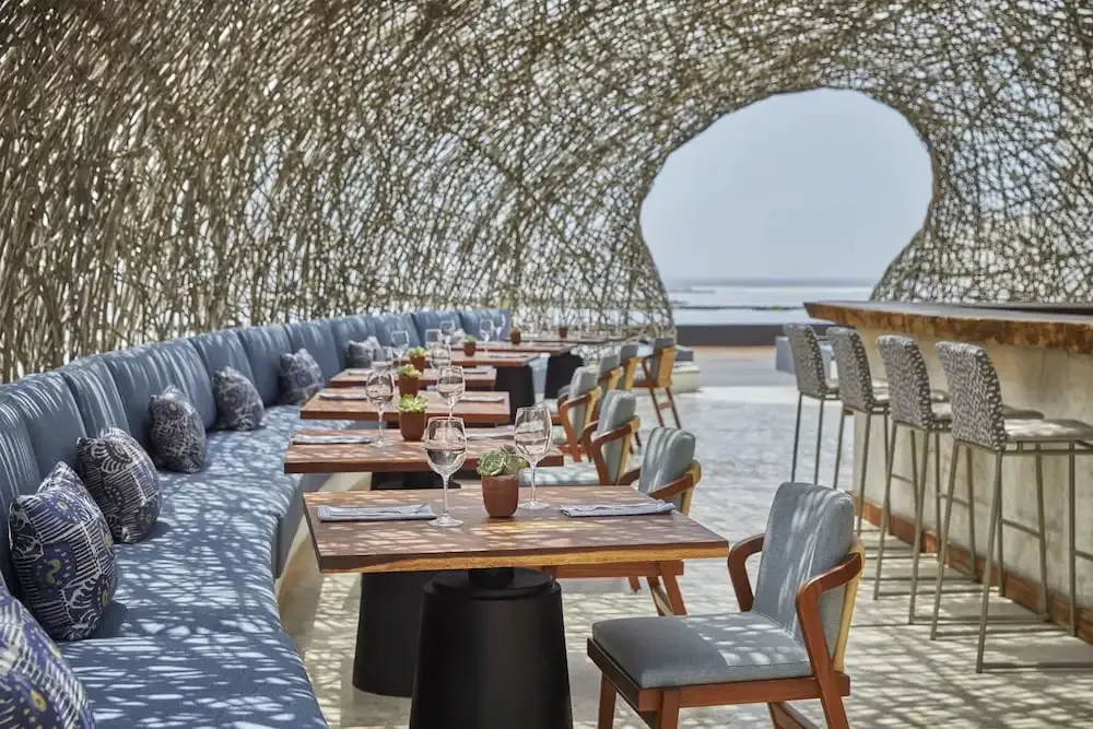 Une salle à manger extérieure avec des tables en bois et des banquettes bleues sous une structure ajourée.