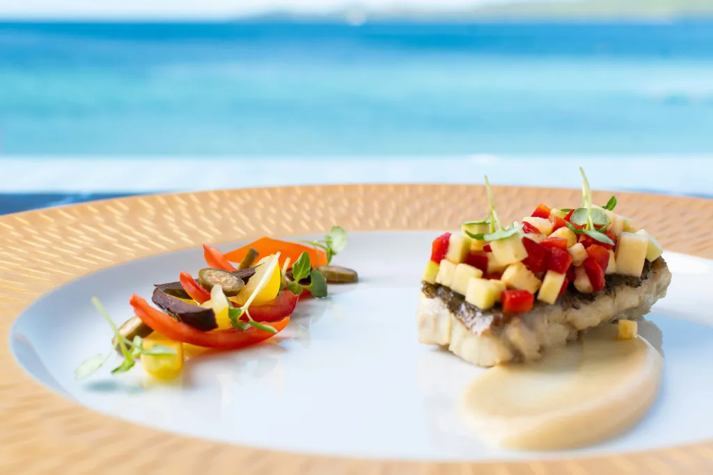 Assiette de poisson frais décorée de légumes et d'herbes, avec vue sur la mer en arrière-plan.