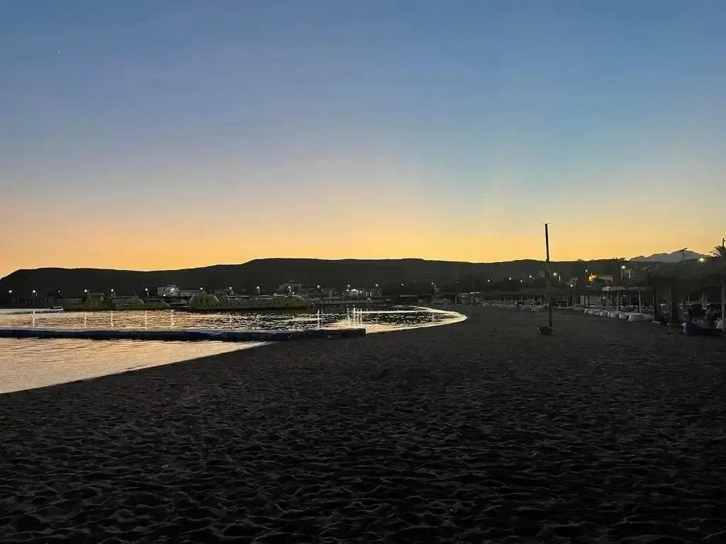 Plage de sable sombre au coucher du soleil, avec des transats alignés.