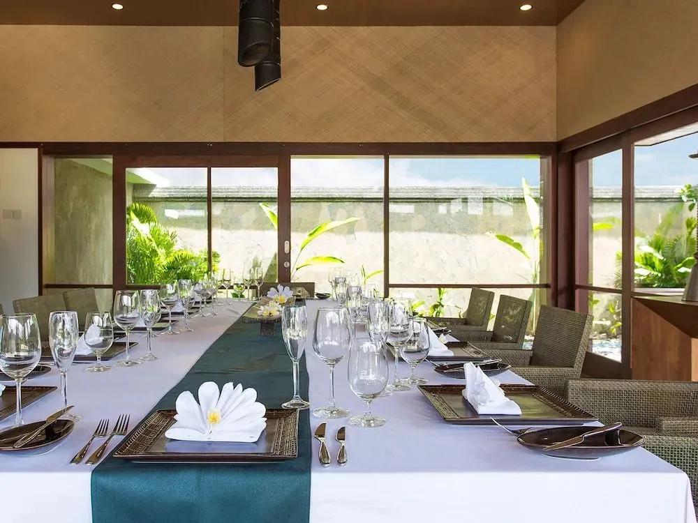 Une longue table est dressée pour un repas dans une salle lumineuse avec vue sur la végétation.
