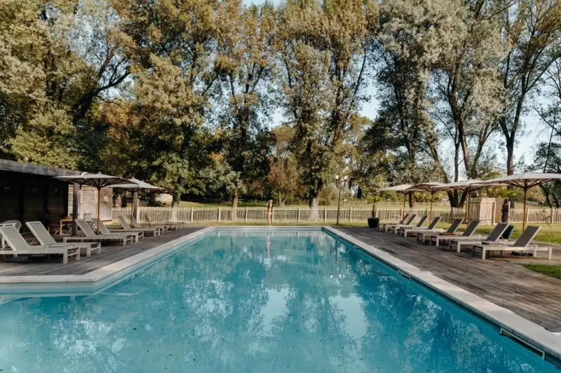 Une piscine rectangulaire bordée de transats sous des parasols, entourée d'arbres.