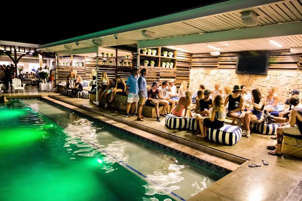 Des personnes sont assises au bord d'une piscine illuminée la nuit.
